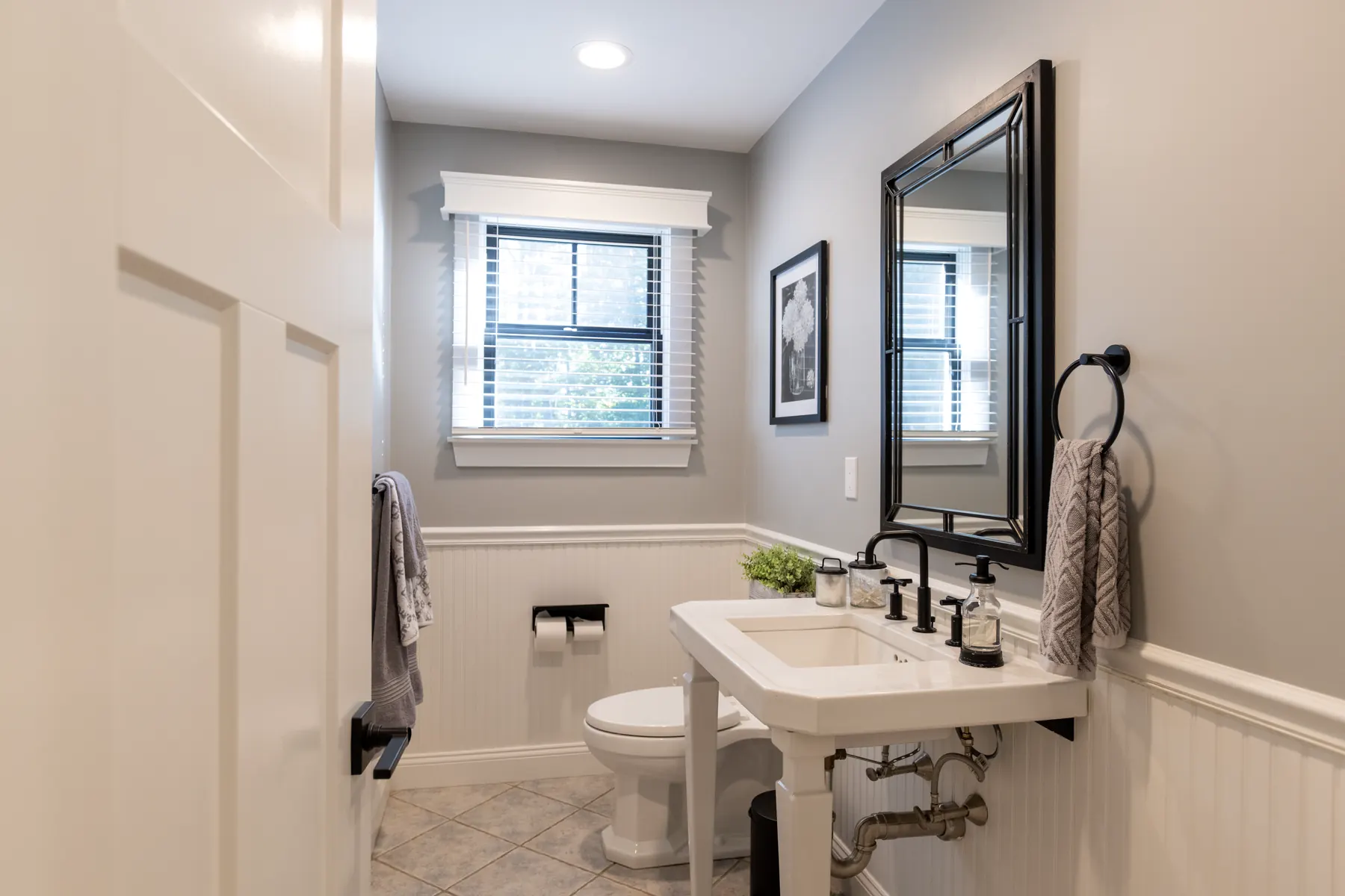 Powder room with a pedestal sink, black-framed mirror, and white wainscoting.