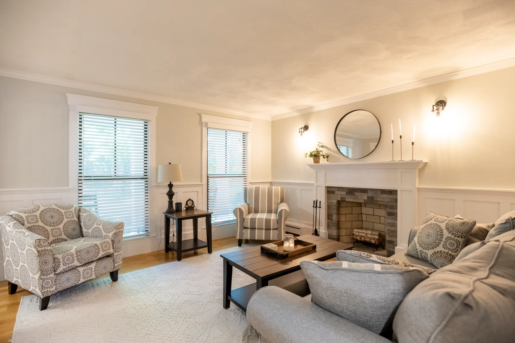 Living room with a white fireplace, patterned chairs, and tall windows.