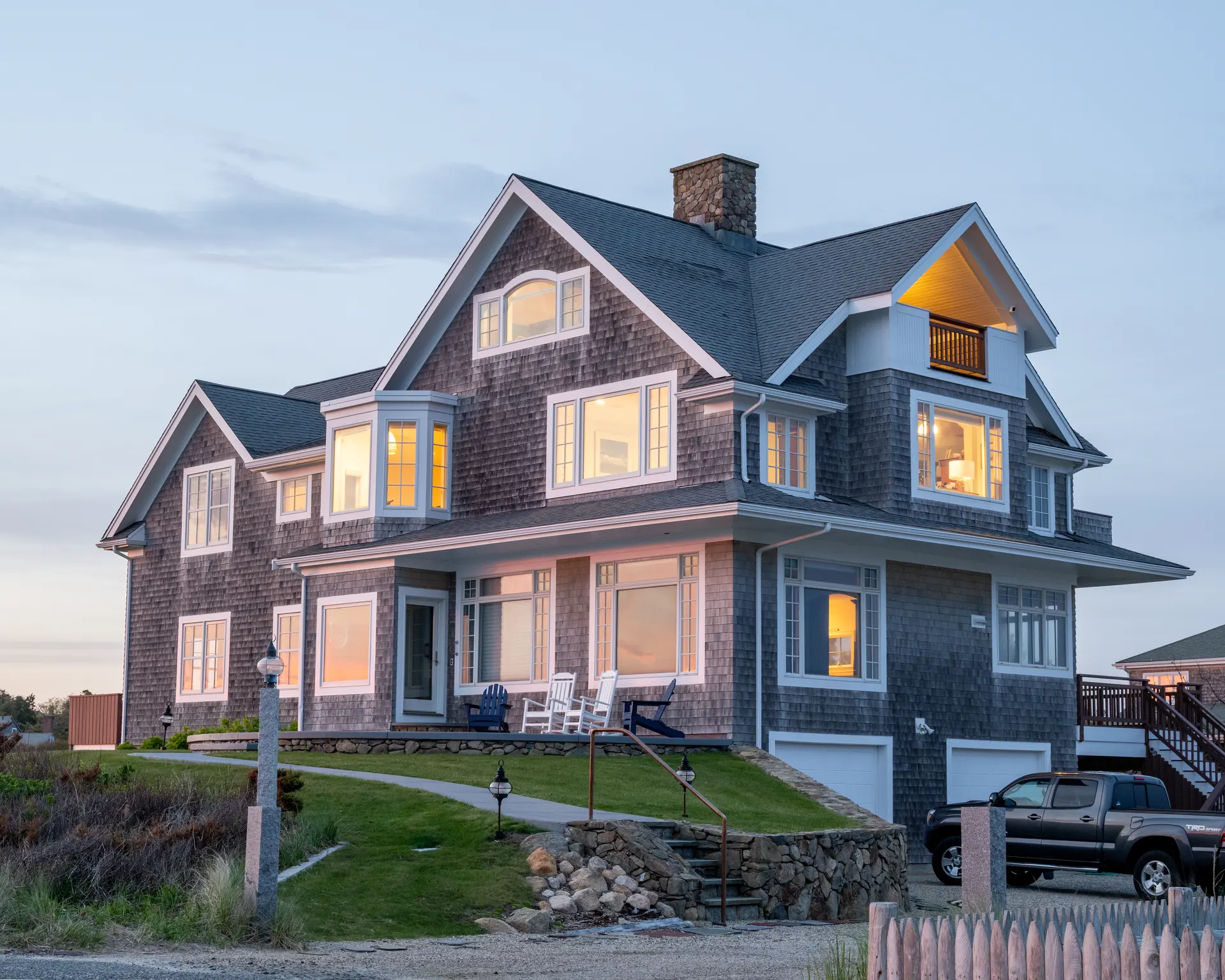 Shingle-style coastal home photographed at dusk with warm interior lights.