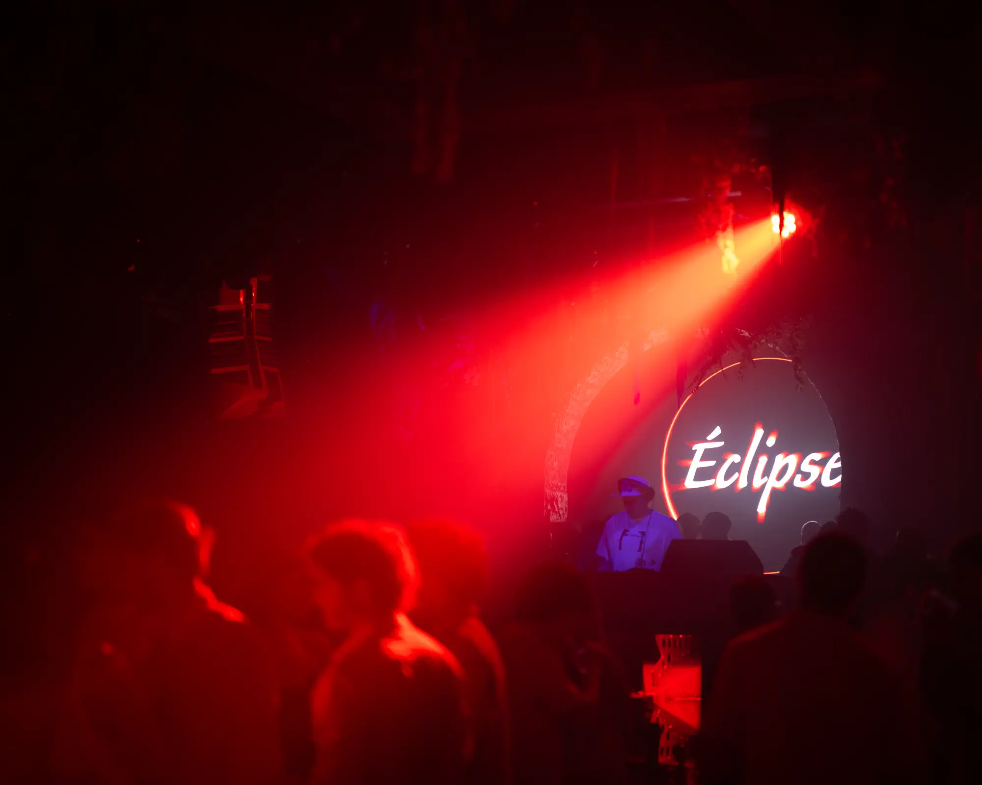 Red stage light cuts through a crowded nightclub toward the Eclipse DJ booth.