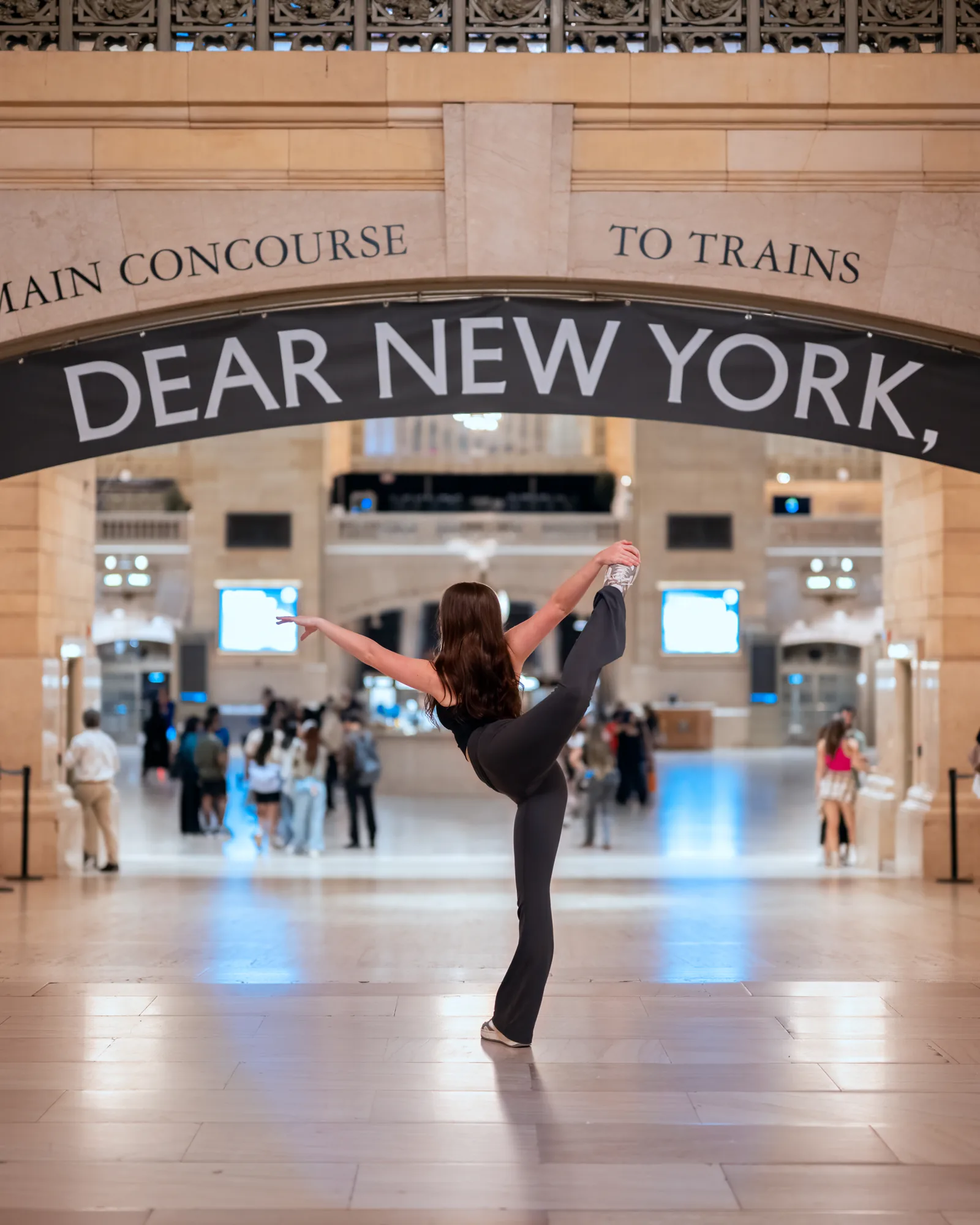Dancer holding an arabesque beneath a Dear New York sign inside a train terminal.