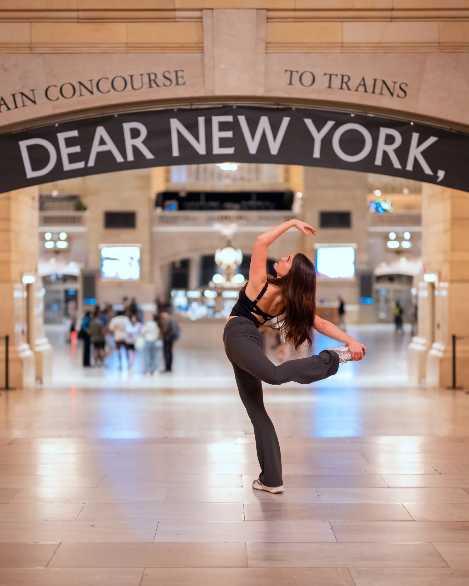 Dancer holding a bent-leg balance beneath a Dear New York sign inside a train terminal.