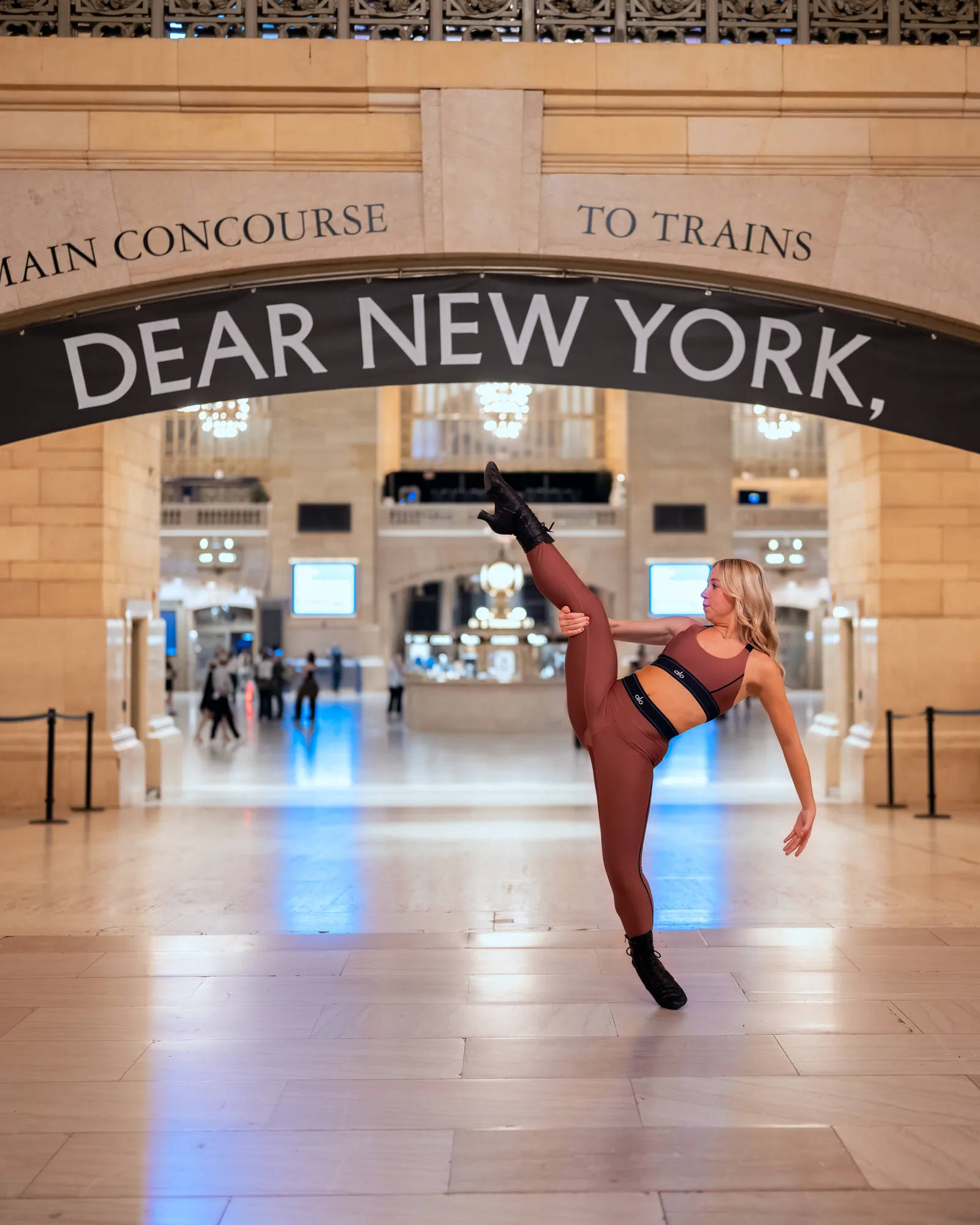 Dancer holding a high leg extension beneath a Dear New York sign inside a train terminal.