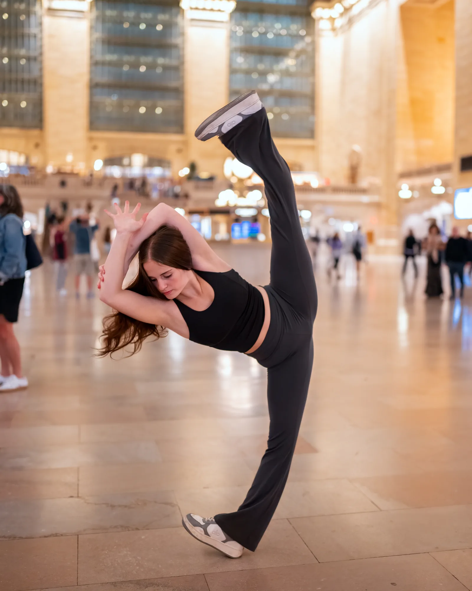 Dancer holding a high leg extension on a New York City train terminal concourse.