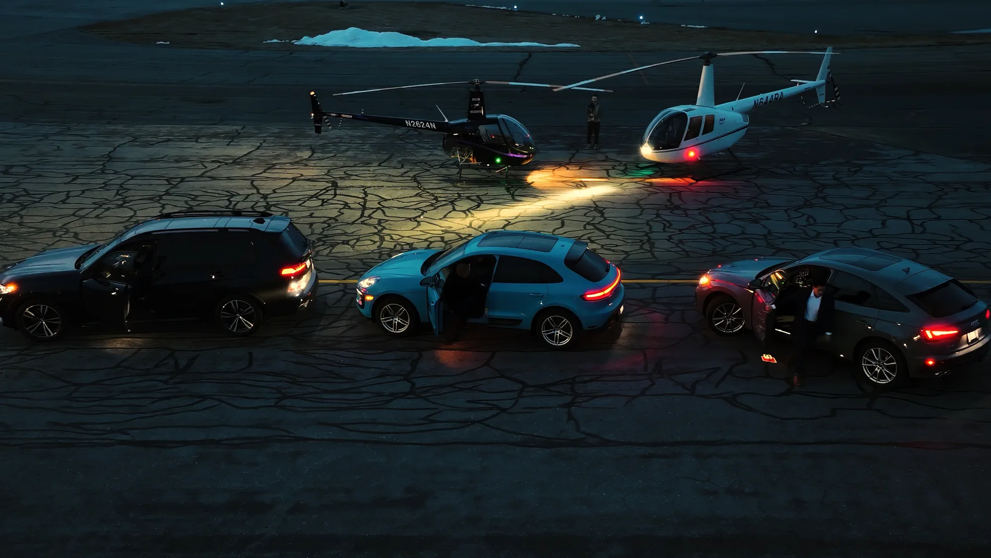 Three vehicles lined up on an airstrip facing two helicopters at dusk.