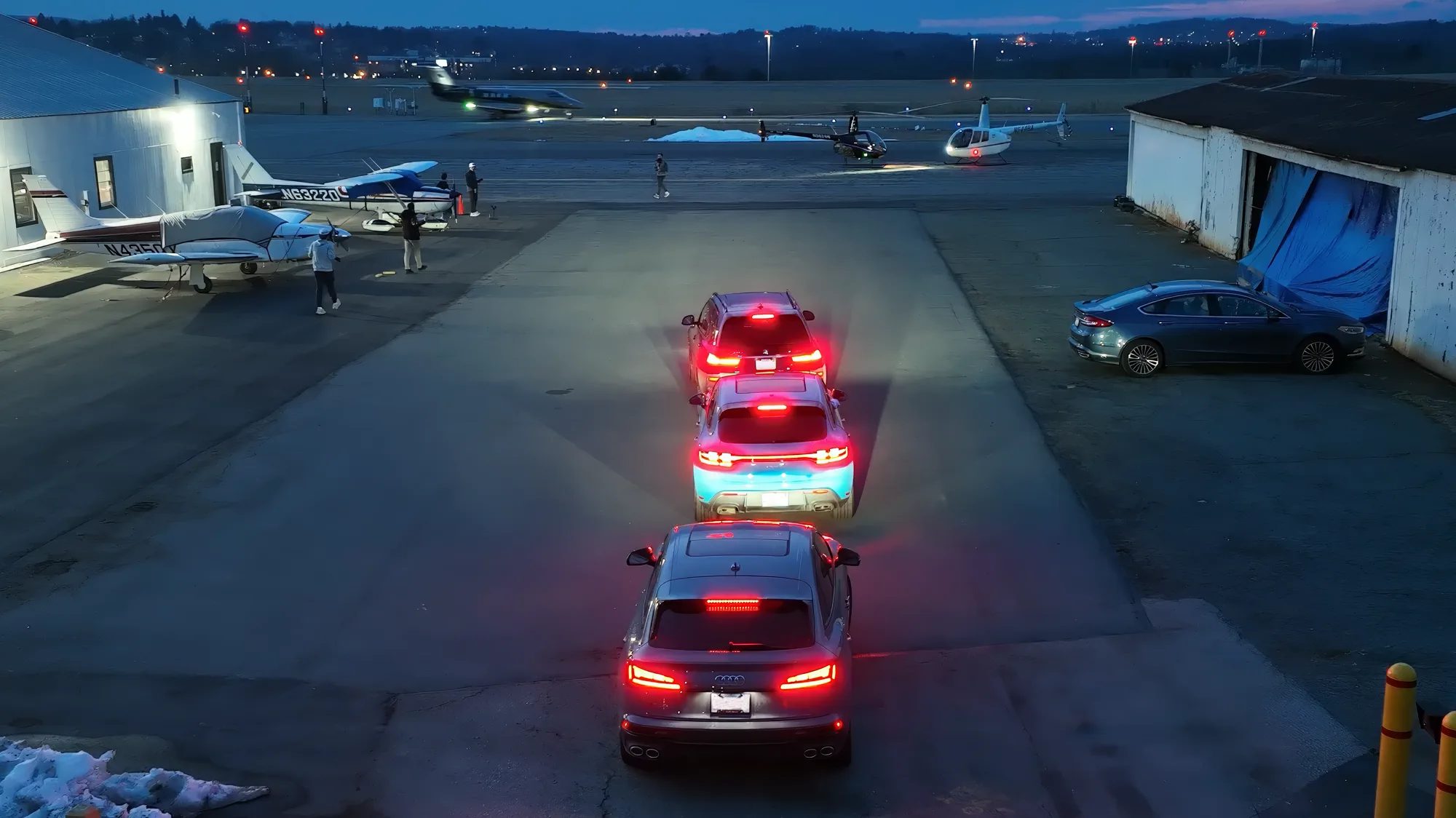 Three-car convoy approaching two helicopters on a dusk airstrip.