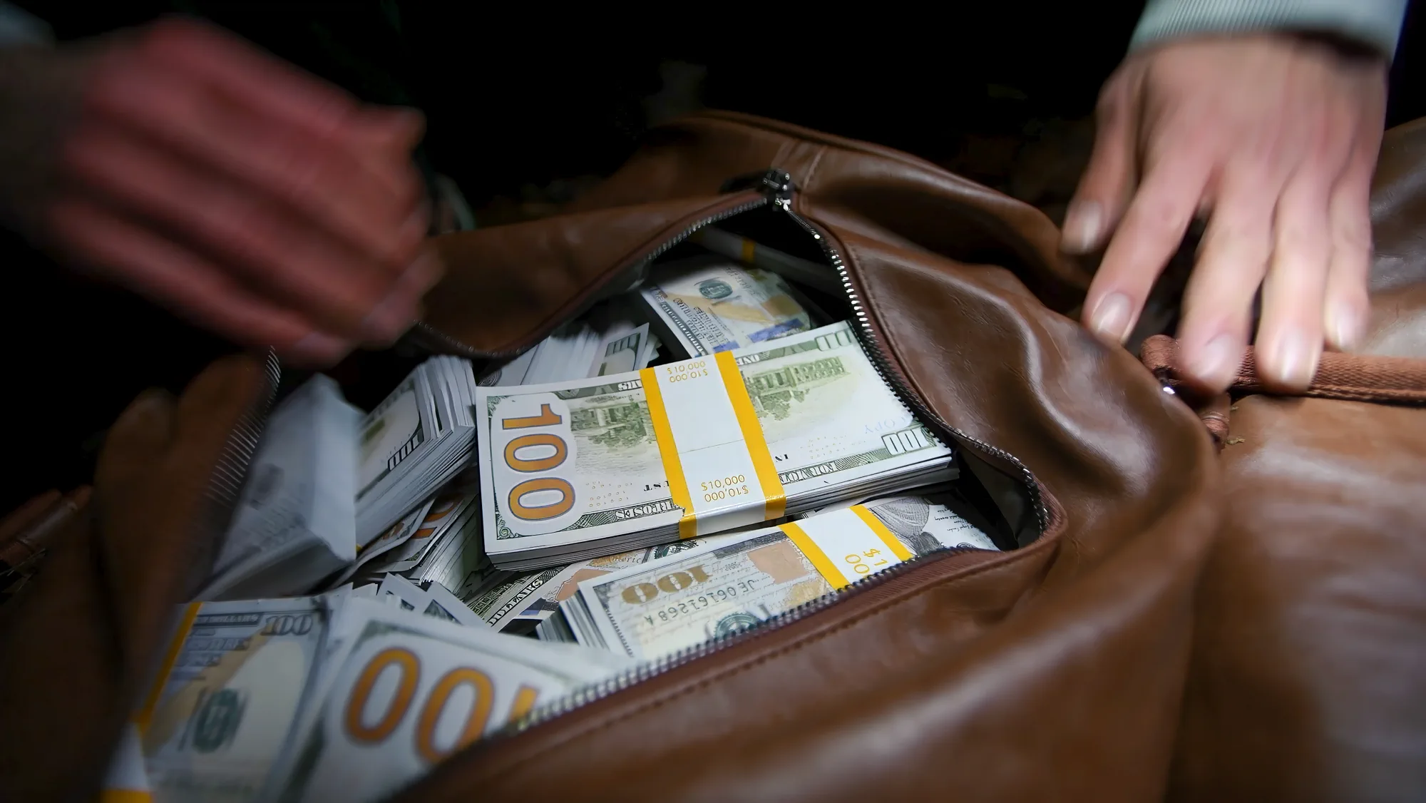 Hands opening a brown duffel bag filled with bundled banknotes.
