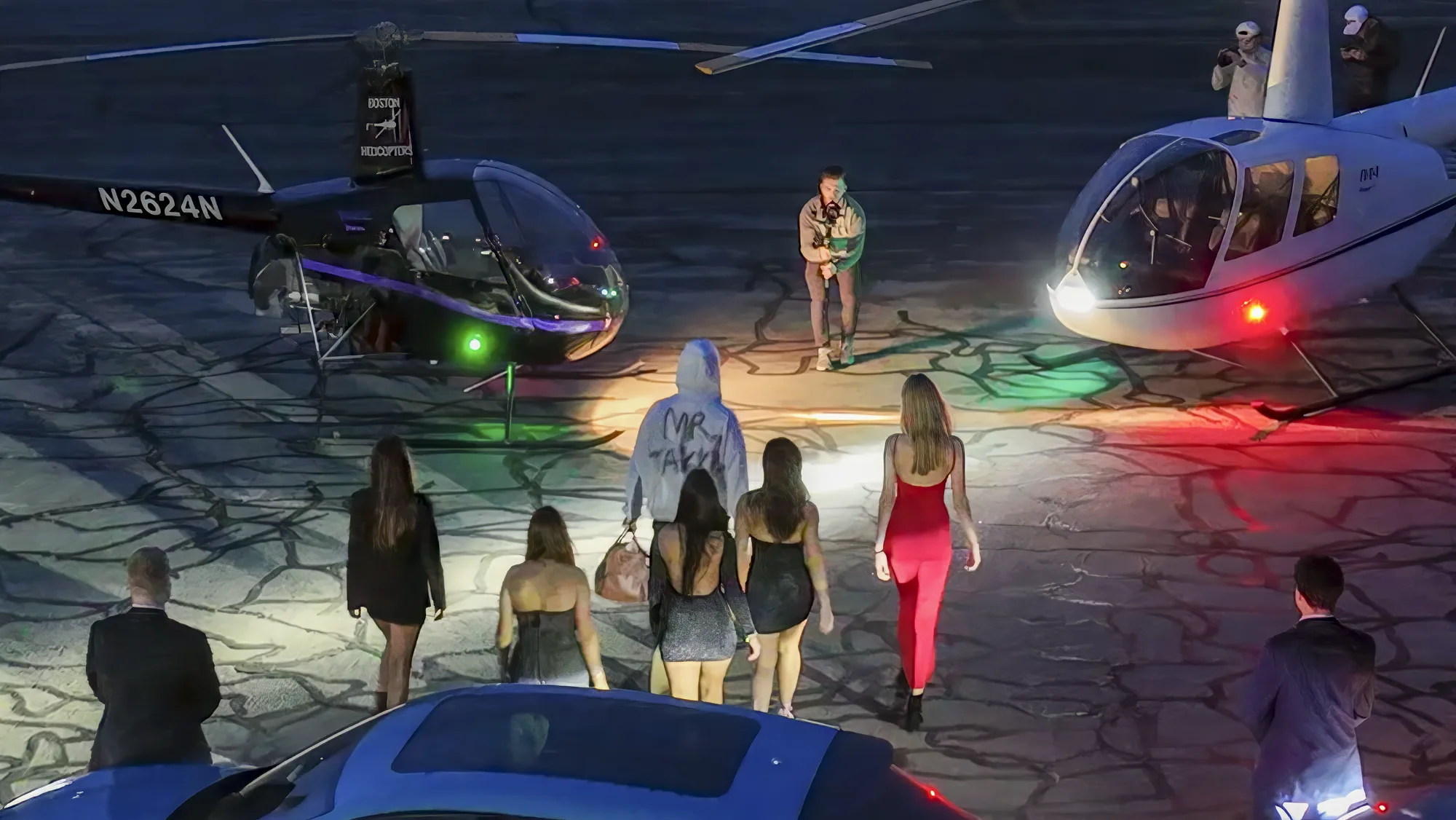 Performer and cast walking with duffel bags toward two illuminated helicopters.