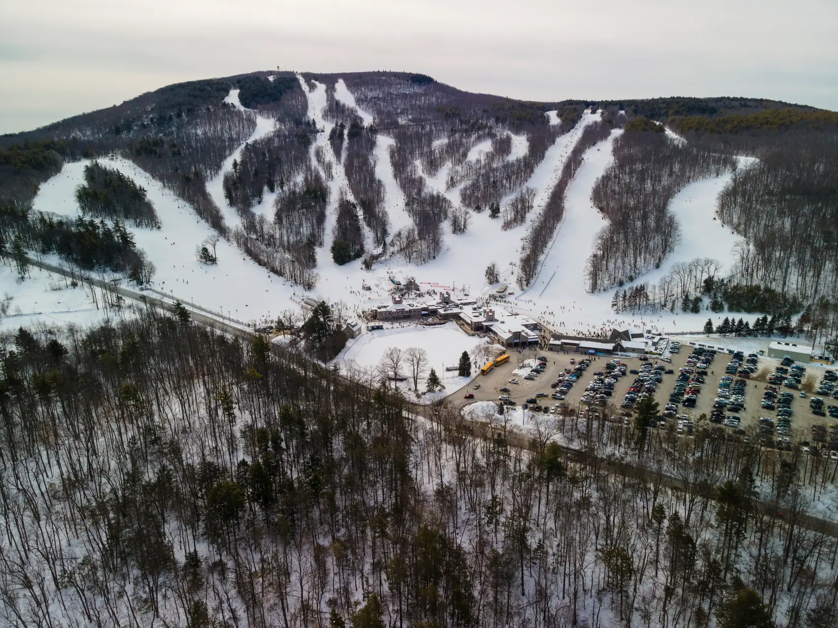 Wide aerial view of a winter resort, surrounding woods, base buildings, and parking area.