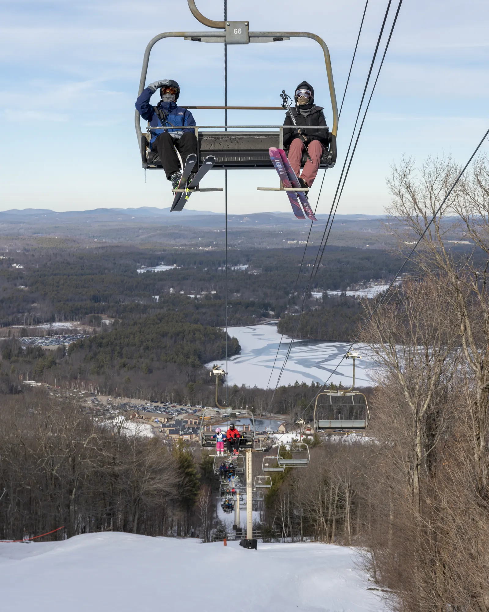 Two skiers riding a chair lift above a snow-covered trail and wooded valley.