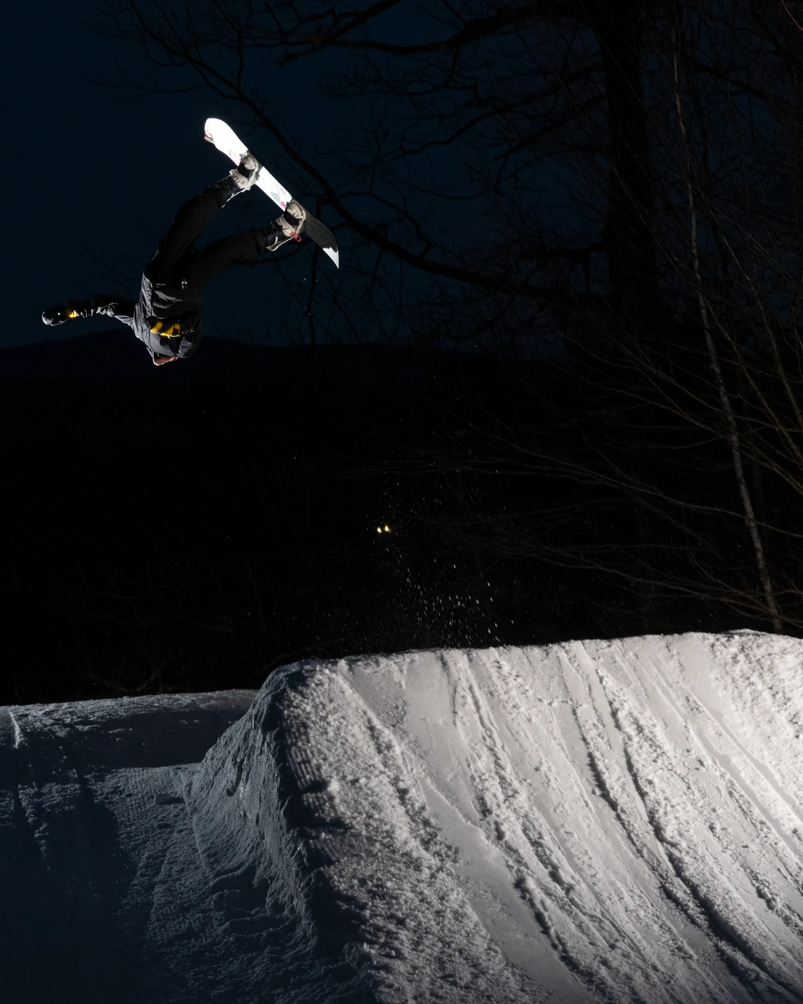 Snowboarder rotating through a backflip above a snow jump at night.
