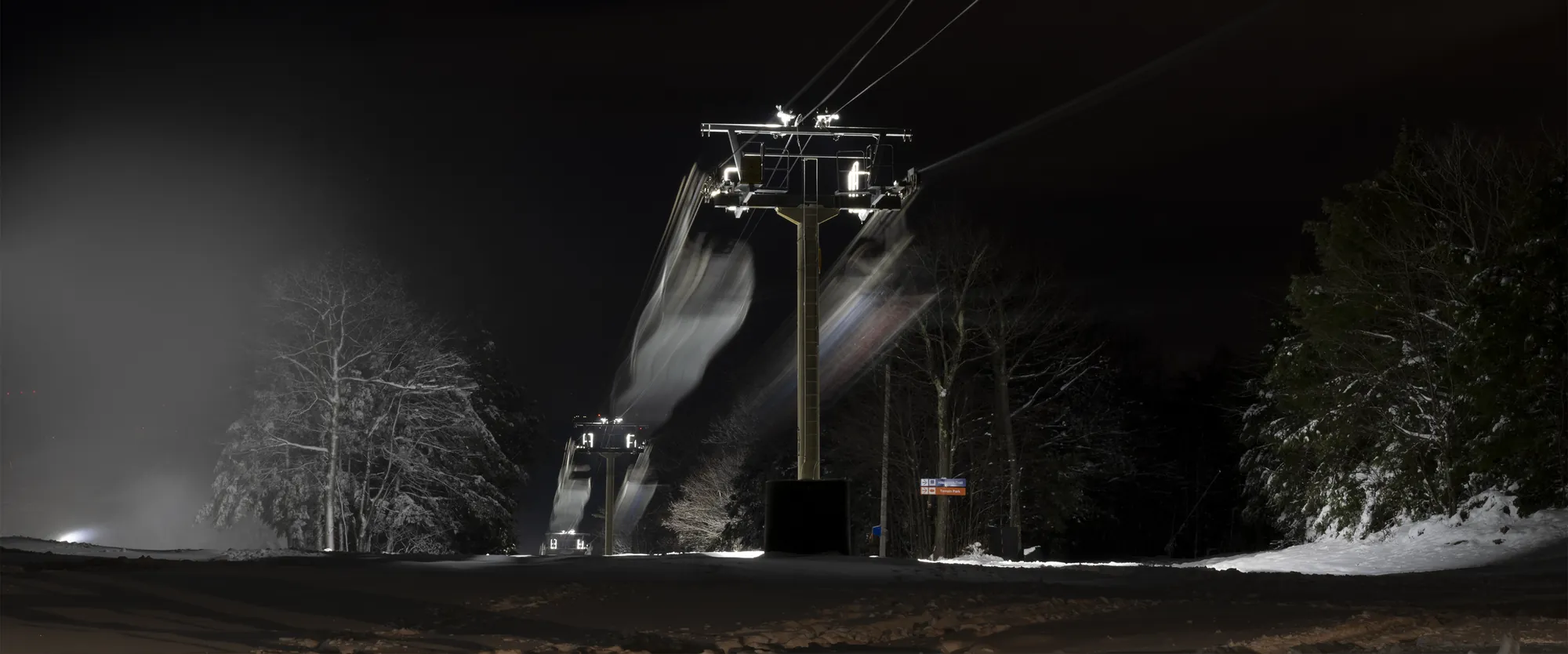 Chair lifts passing a floodlit snowmaking plume after dark.